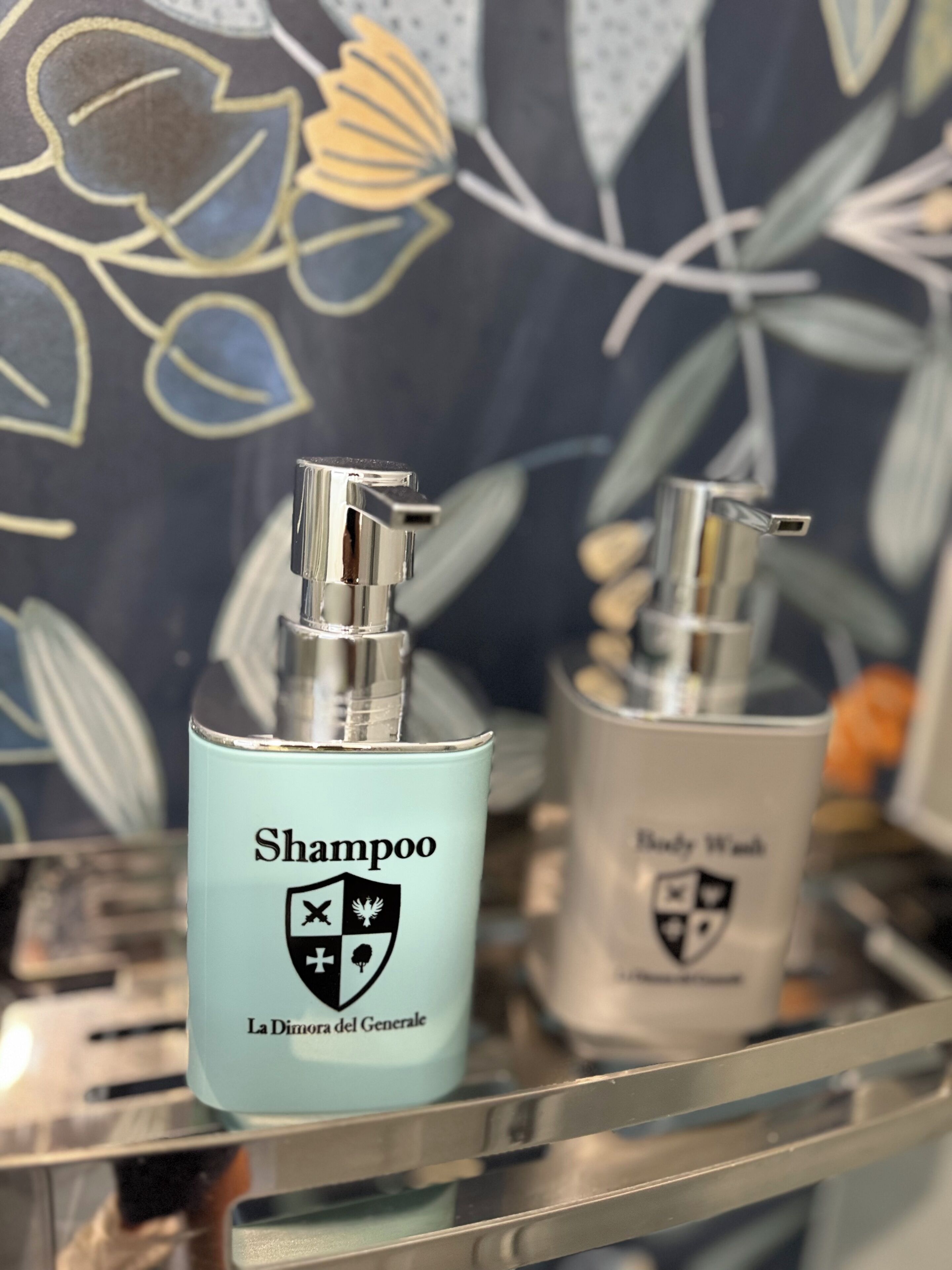 Twee shampoo flessen op een metalen plank, met een bloemenpatroon op de achtergrond.