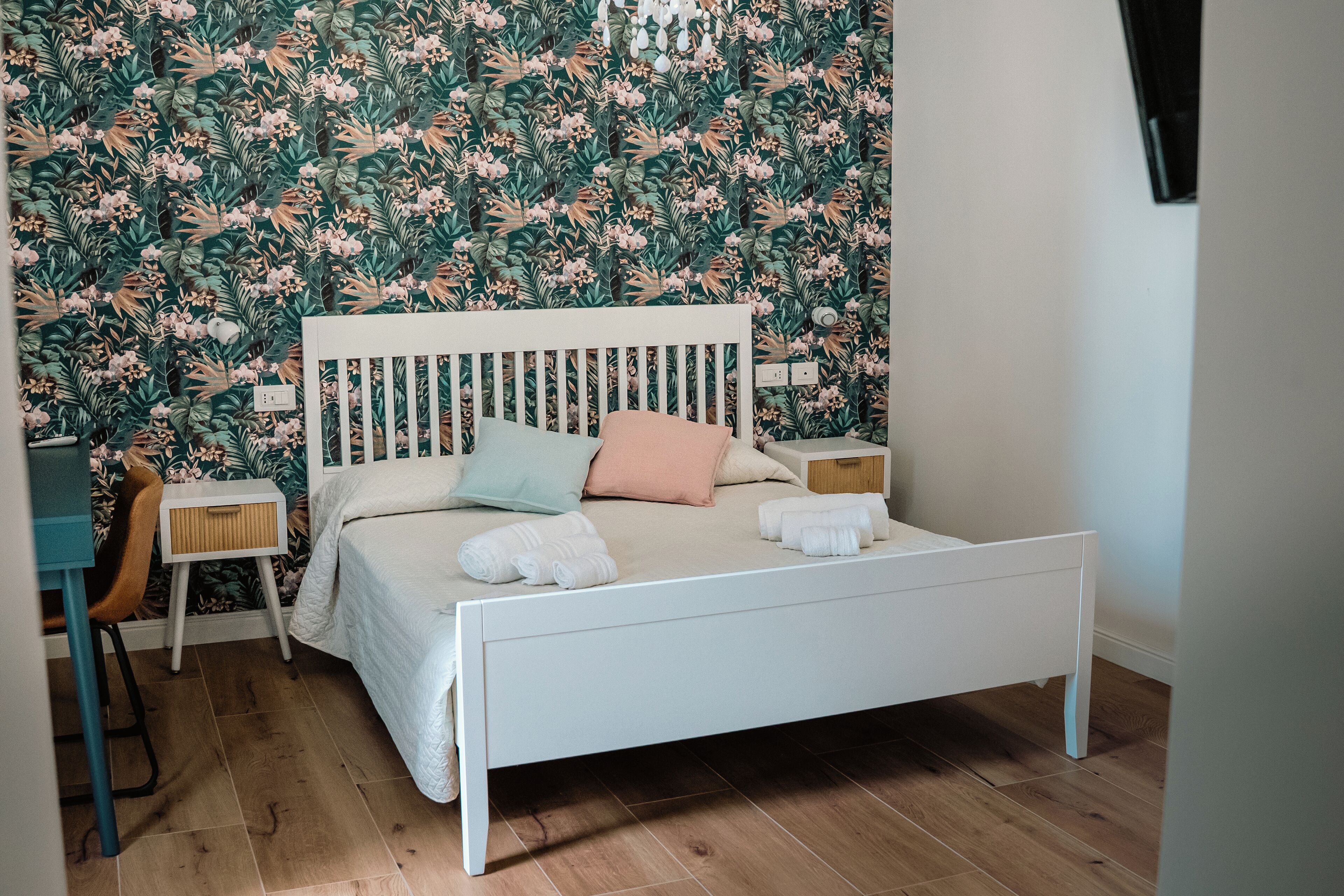 Un dormitorio con una cama blanca, una mesita de noche pequeña, una silla y una pared con papel tapiz floral.
