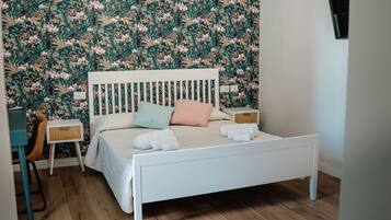 Un dormitorio con una cama blanca, una mesita de noche pequeña, una silla y una pared con papel tapiz floral.