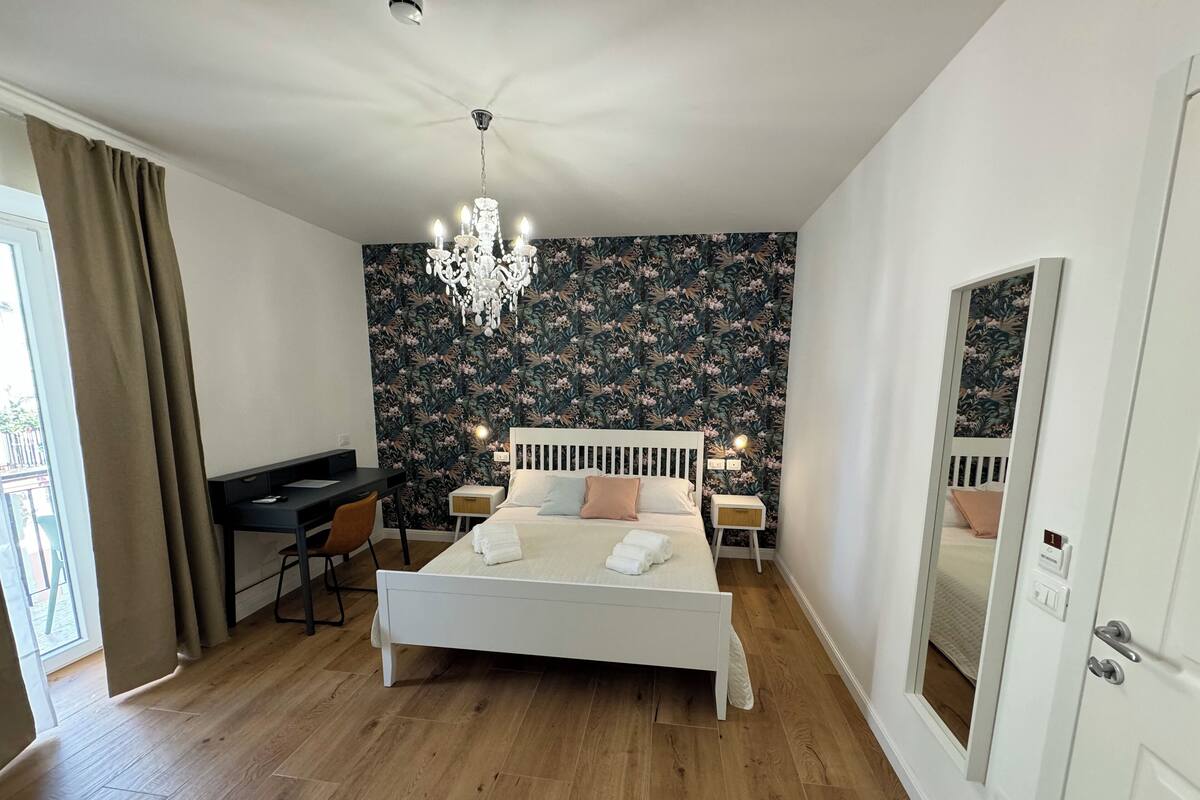 A bedroom with a bed, a desk, a chair, a chandelier, and a mirror.