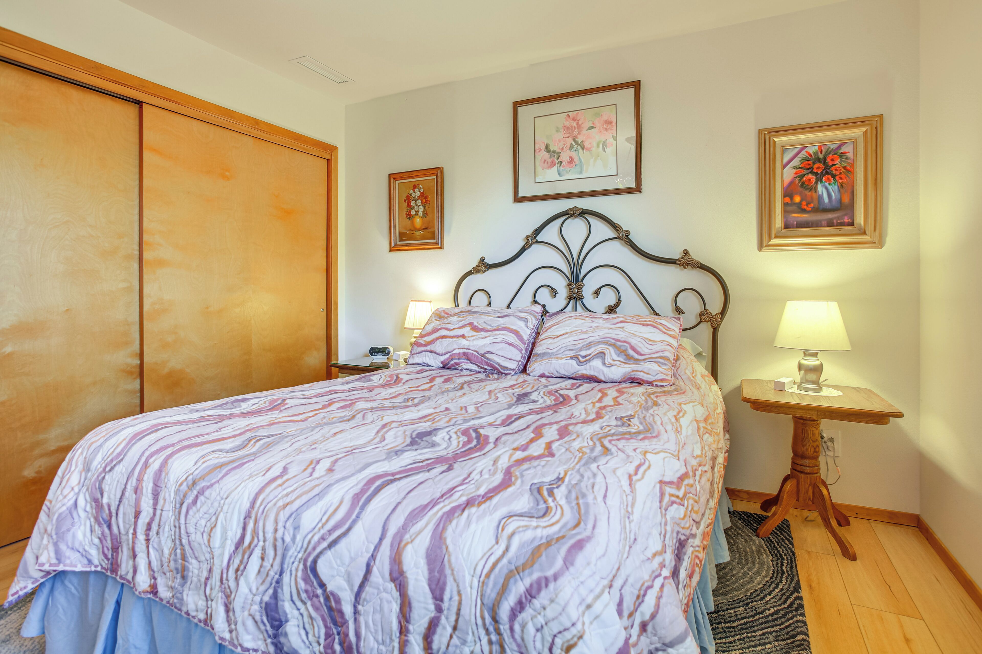 A bedroom with a bed, two bedside tables with lamps, a wooden wardrobe, and two framed floral paintings on the wall.