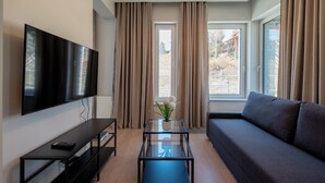 Una sala de estar moderna con un televisor de pantalla plana, una mesa de centro de vidrio, un sofá y ventanas grandes con cortinas.
