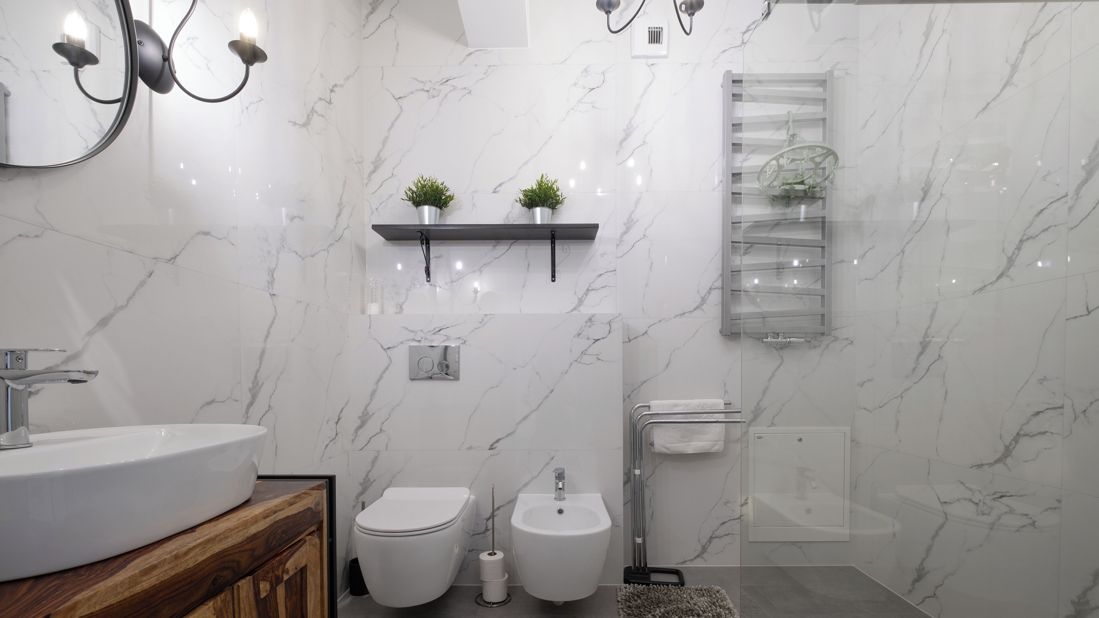 A modern bathroom with a marble backsplash, a wooden vanity, a toilet, a bidet, and a glass shower enclosure.