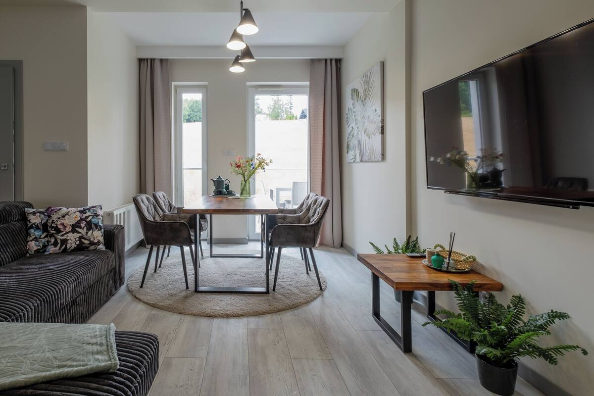 Un salon moderne avec un canapé, une table à manger et des chaises, un téléviseur fixé au mur et une plante en pot.