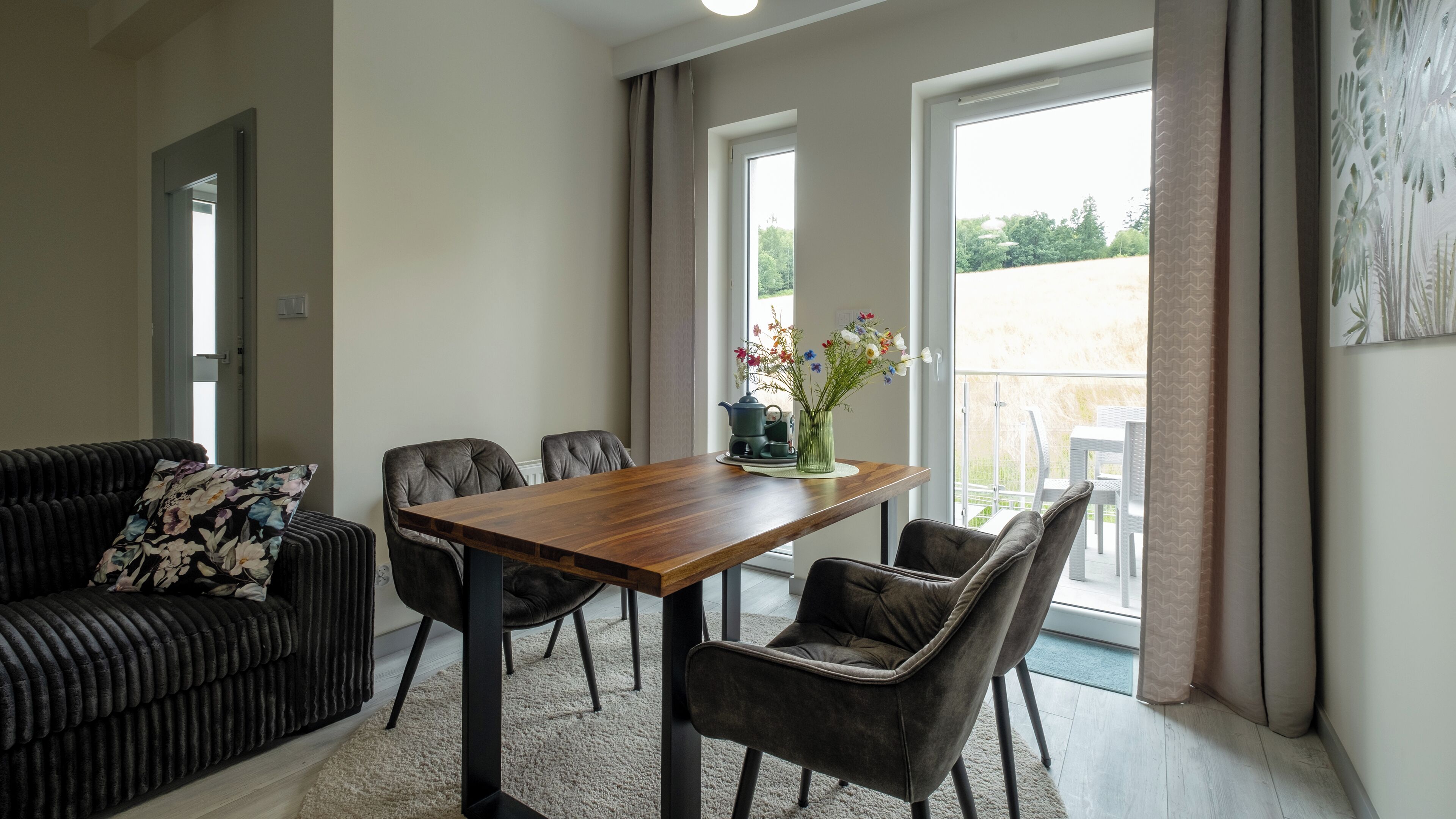 A modern living room with a wooden dining table, two grey upholstered chairs, a black sofa with a floral cushion, and a large window with a view of greenery.