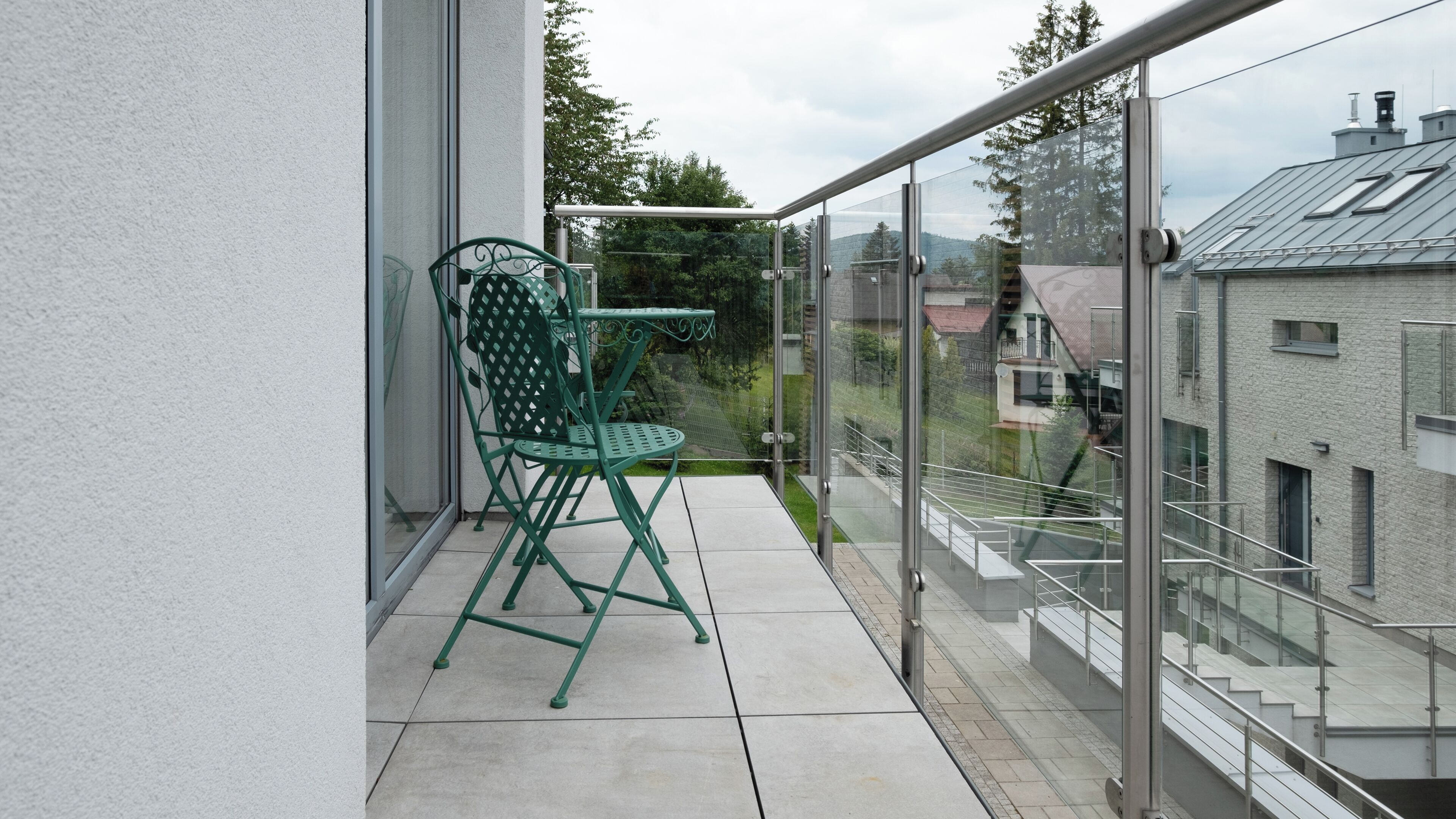 En balkon med en grøn metalstol og et bord, glasgelændere samt udsigt til bygninger og træer.
