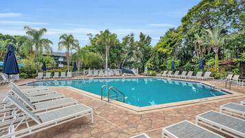 A rectangular swimming pool surrounded by lounge chairs and umbrellas, with a tiled deck and tropical vegetation.