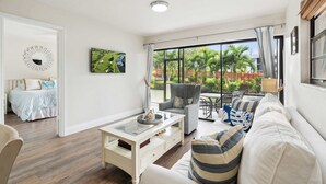 A living room with a sofa, armchair, coffee table, and a dining area with a table and chairs. There is a large window with curtains, a painting on the wall, and a bed in the adjacent room. - B-116 - Resort Views and Close to the Lobby (Naples)
