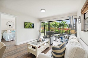 Ein Wohnzimmer mit einer Couch, Sesseln, einem Couchtisch und einem Essbereich mit Tisch und Stühlen. Ein großes Fenster mit Vorhängen, ein Gemälde an der Wand und ein Bett im angrenzenden Zimmer.