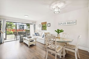 A spacious living room with a glass dining table, white sofas, and a large sliding glass door leading to an outdoor area. - B-116 - Resort Views and Close to the Lobby (Naples)