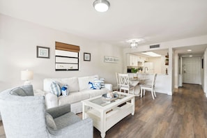 A modern living room with a white sofa, a grey armchair, and a coffee table. There is a dining area with a white table and chairs. The kitchen is visible in the background with white cabinets. - B-116 - Resort Views and Close to the Lobby (Naples)