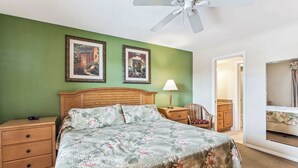 Un dormitorio con cama de madera, paredes verdes, dos cuadros enmarcados, una mesita de noche, una lámpara, una silla y un ventilador de techo.