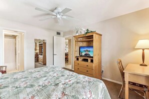 A bedroom with a bed, a TV on a wooden stand, a ceiling fan, and a desk with a lamp.