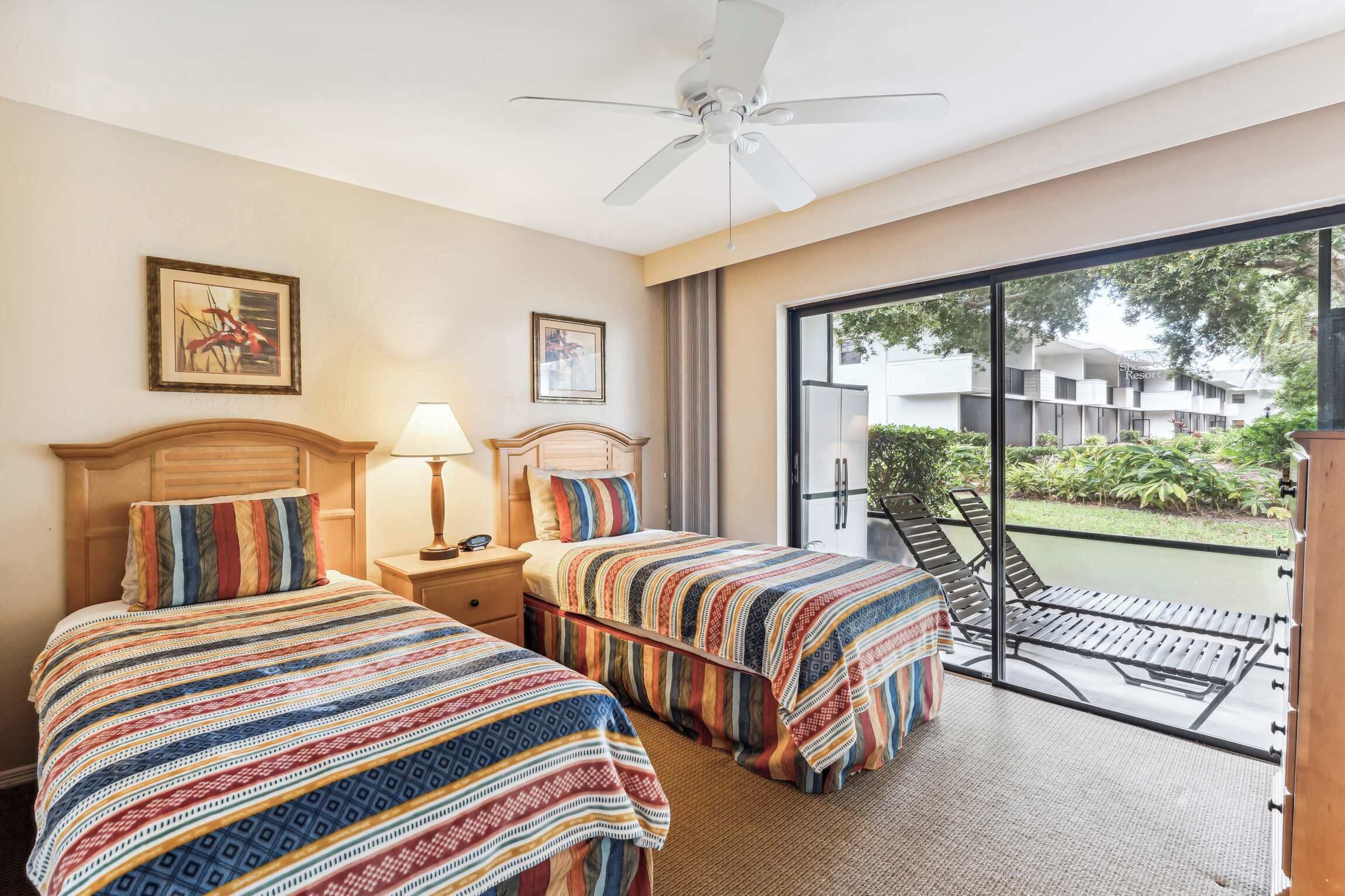 A hotel room with two beds, a ceiling fan, a nightstand, a lamp, and a view of the outside.