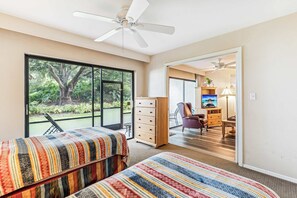 A bedroom with two beds, a ceiling fan, a large window with a view of greenery, and a door leading to a bathroom with a shower and a toilet. - D-126 Resort Views, Quiet Back End of Resort (Naples)