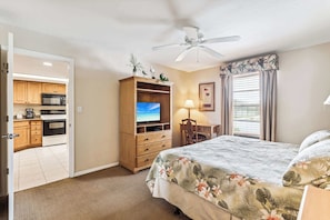 A hotel room with a bed, a TV, a ceiling fan, a desk, and a view of the kitchen.