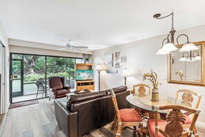 A modern living room with a glass door leading to a balcony, a leather sofa, a dining table with chairs, and a ceiling fan.