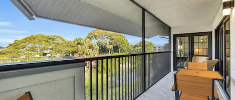 A balcony with a view of a green landscape, a table with chairs, and a glass door leading to an interior space.