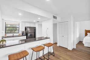 A modern kitchen with a black countertop, white cabinets, and a stainless steel refrigerator. There are two stools at the island and a wooden door leading to a bedroom.