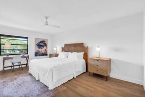A bedroom with a wooden bed, two bedside tables with lamps, a desk with a chair, and a large window with a view of greenery.