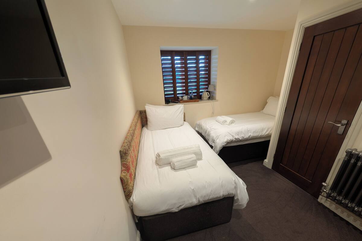 A small hotel room with two single beds, a window with blinds, a flat-screen TV mounted on the wall, and a door with a handle.