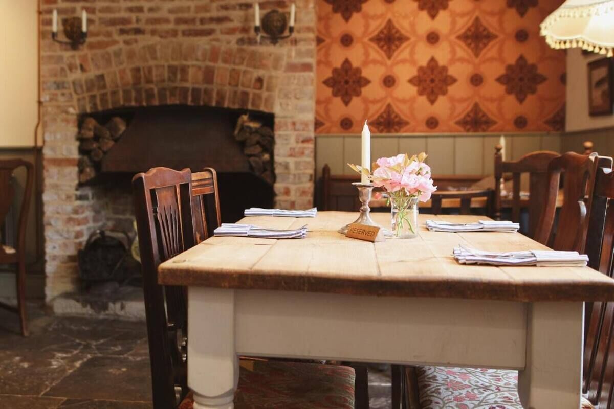A dining area with a brick fireplace, a table set with papers and a candle, and patterned wallpaper.