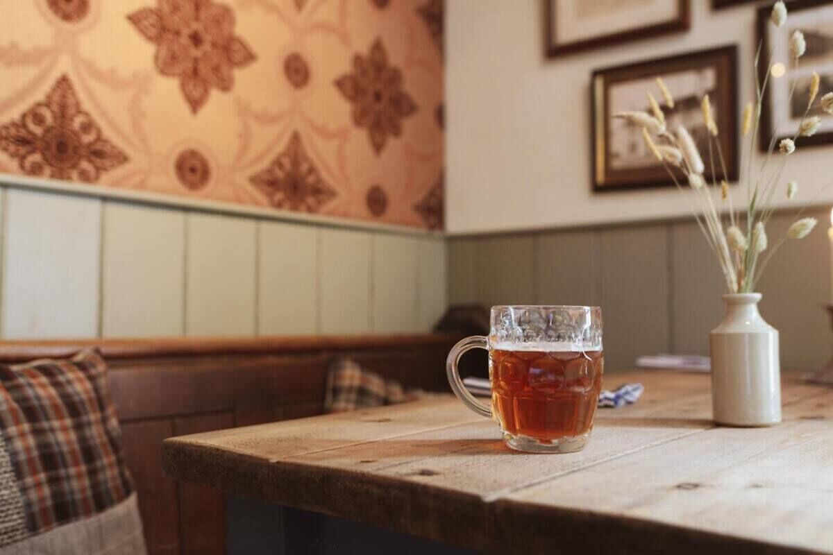 Una mesa de madera con una taza de vidrio de té, un jarrón con plantas secas y un papel tapiz estampado al fondo.