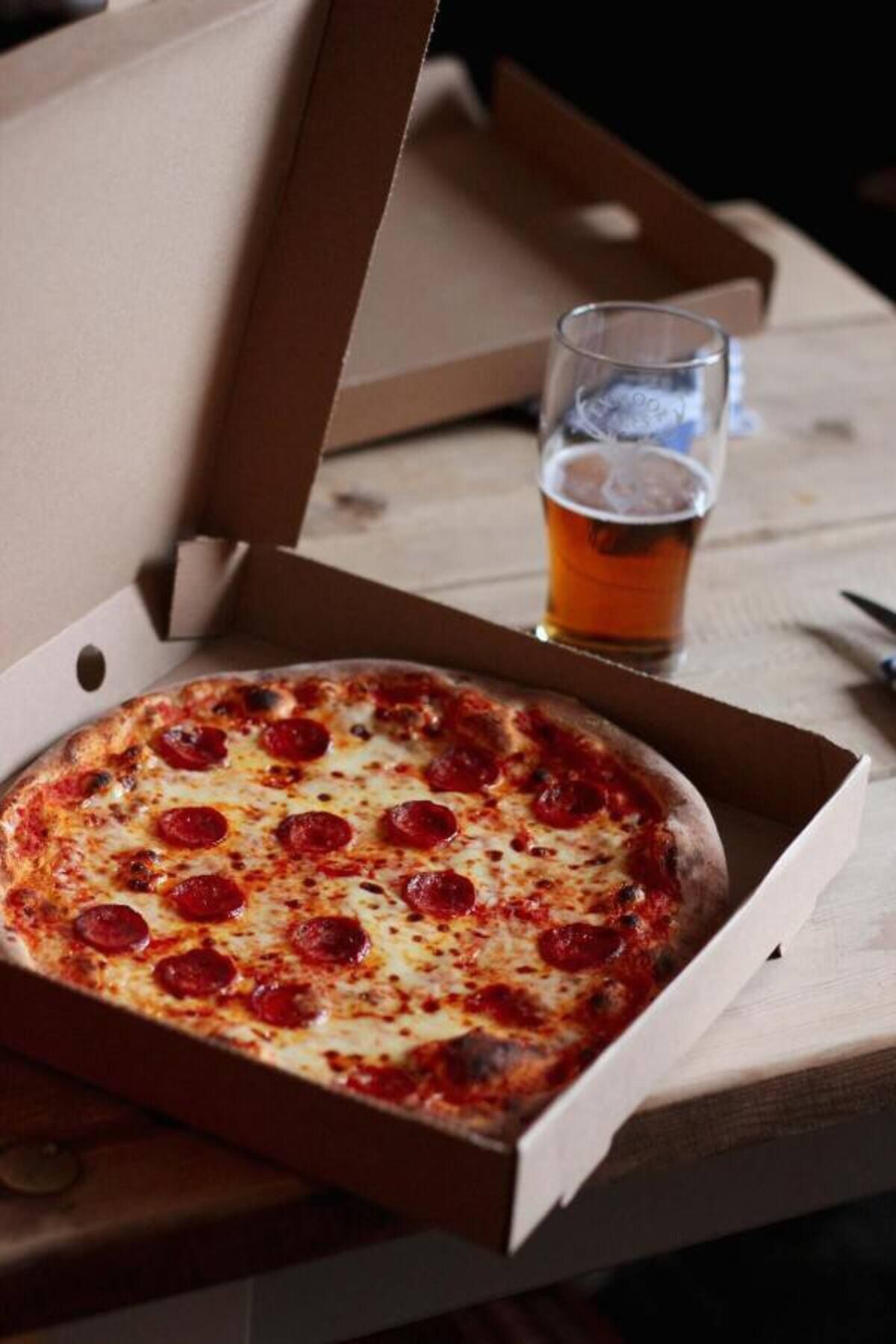 A pizza in a cardboard box on a wooden table with a glass of beer beside it.
