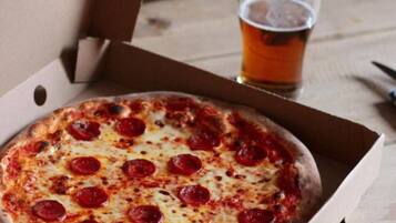 A pizza in a cardboard box on a wooden table with a glass of beer beside it.
