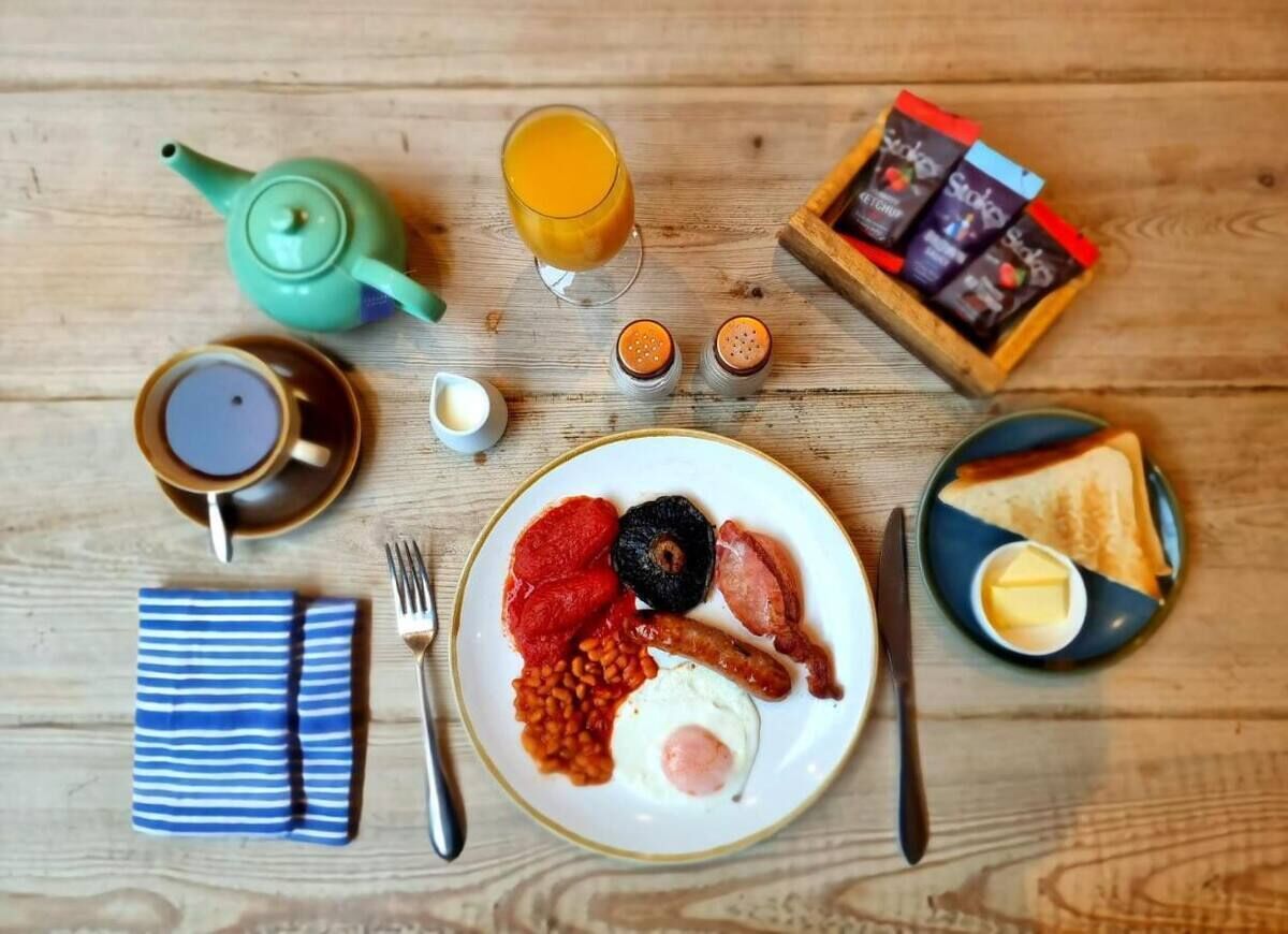 Un desayuno con tetera, taza, tostadas, mermelada, mantequilla y un plato de huevos, frijoles y salchichas.