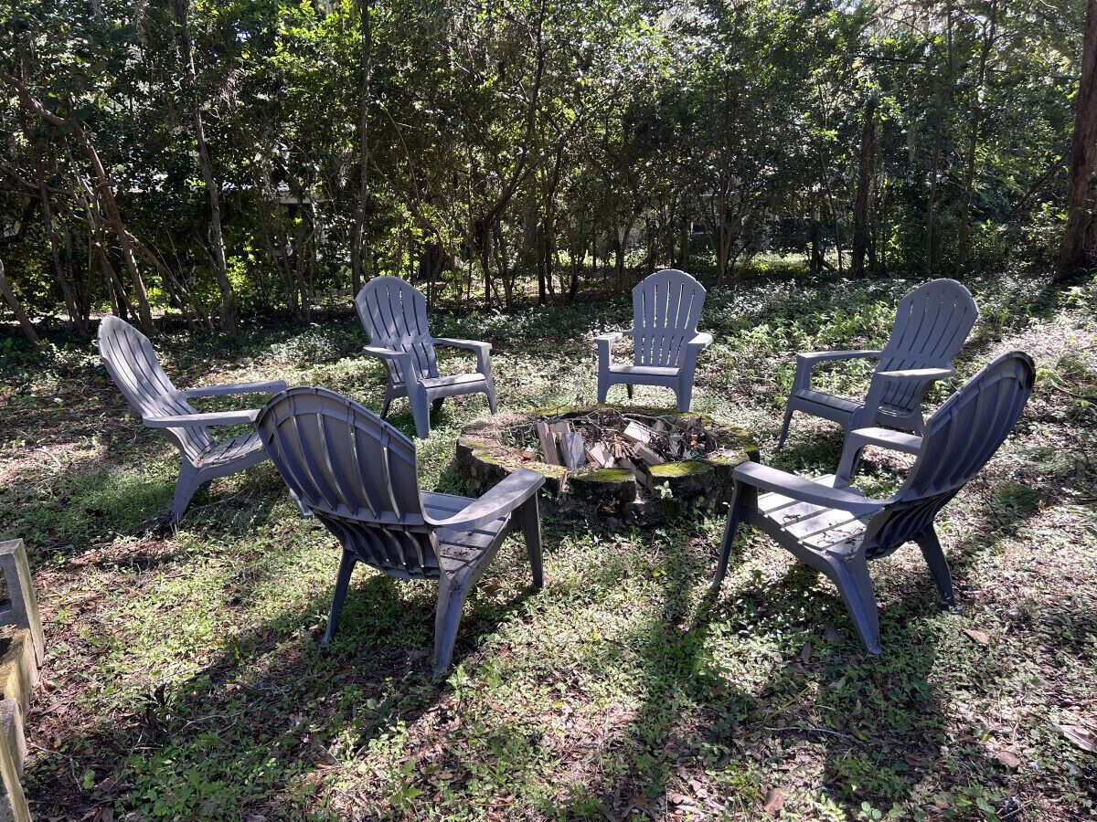 A group of Adirondack chairs arranged around a fire pit in a wooded area.