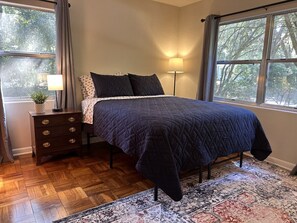 A bedroom with a bed, a nightstand with drawers, a lamp, and a window with a view of trees.