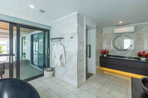 A modern bathroom with a glass shower enclosure, a marble-tiled floor, a dark-toned vanity with a round mirror, and a white robe hanging on a rack.