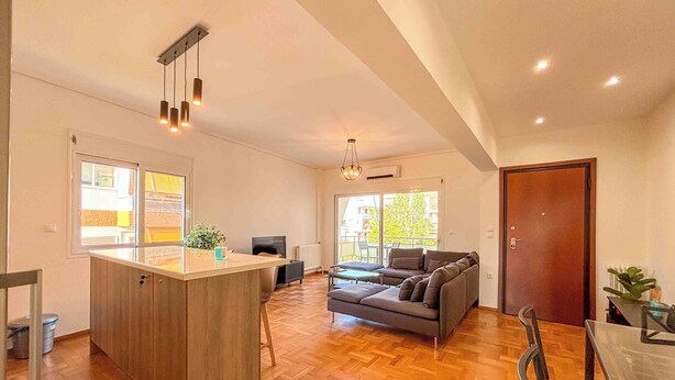 A spacious living area with a wooden herringbone floor, a central island with a sink, a sofa, and a dining table. - Light & Ample Flat near Glyfada Center (Glifada)