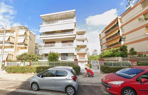 Un edificio residencial de varios pisos con balcones, entrada con portón y automóviles estacionados.