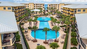 A resort with a central swimming pool surrounded by palm trees and lounge chairs, connected to multi-story buildings with balconies.
