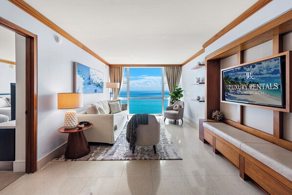 Amplia sala de estar con una gran pantalla de televisión mostrando 'LUXURY RENTALS MIAMI BEACH', un sofá cómodo, una mesa de centro y vista al océano.