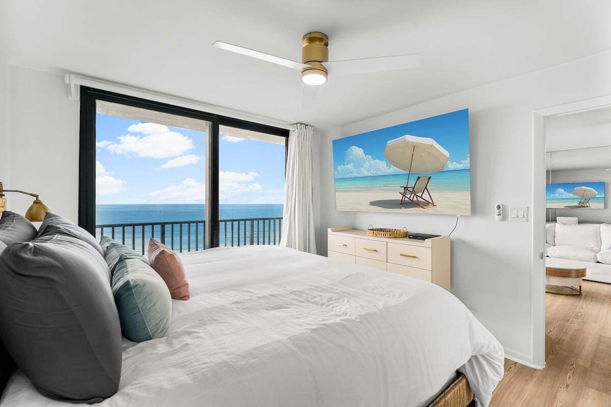 Une chambre avec un grand lit, un ventilateur de plafond, un tableau représentant une scène de plage et une vue sur l’océan par une grande fenêtre.