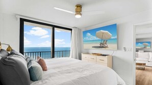 Une chambre avec un grand lit, un ventilateur de plafond, un tableau représentant une scène de plage et une vue sur l’océan par une grande fenêtre.