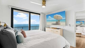 Une chambre avec un grand lit, un ventilateur de plafond, un tableau représentant une scène de plage et une vue sur l’océan par une grande fenêtre.