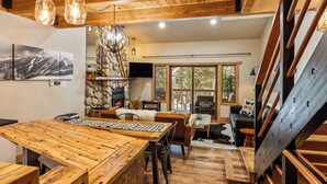 A wooden dining table with chairs, a stone fireplace, and a large window with a view of trees.