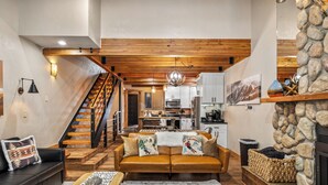 A spacious living area with a wooden staircase, a kitchen in the background, and a stone fireplace.