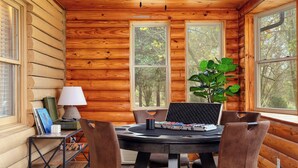 Interior de una cabaña de madera con una mesa redonda, cuatro sillas, una computadora portátil, una lámpara y una planta.