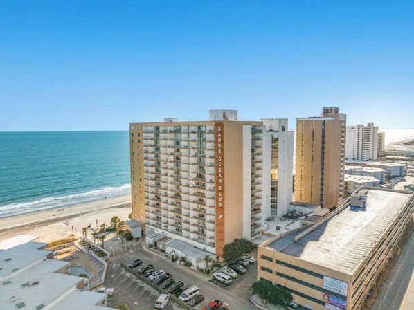 A high-rise hotel with multiple balconies, a beach view, and a parking area.