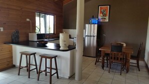 A compact kitchen with a refrigerator, microwave, and a coffee maker. There are two stools and a wooden dining table with chairs. - Woodstock 3 Francis St, Port Elliot (Port Elliot)