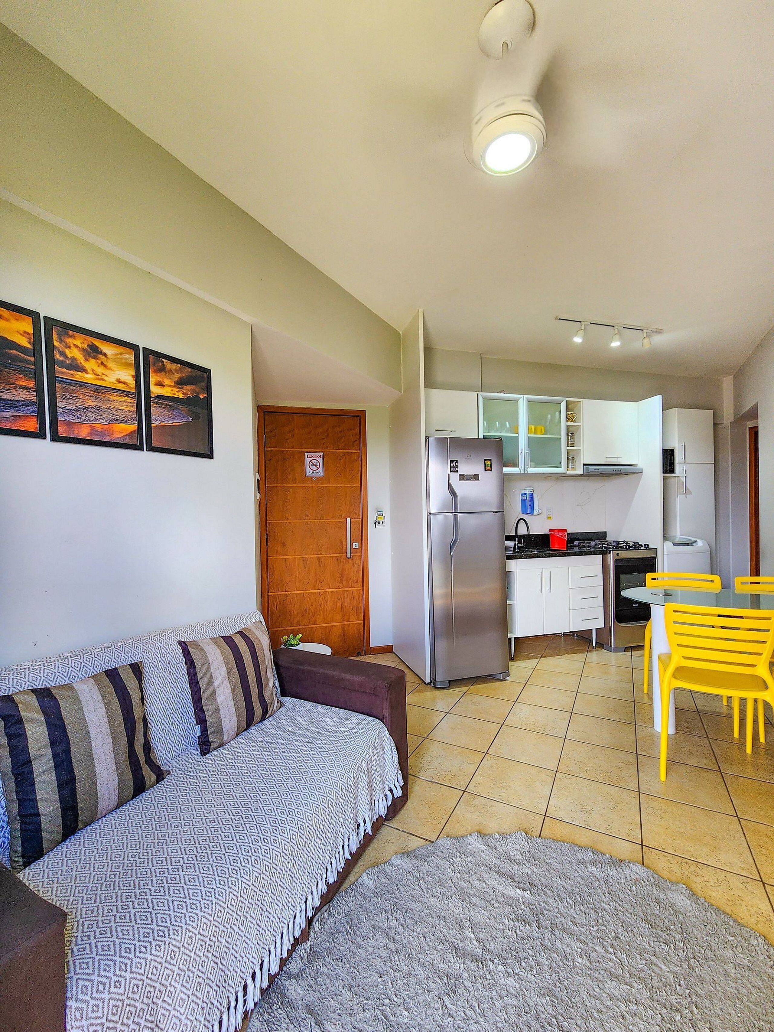 A compact living space with a sofa, a dining table with yellow chairs, a kitchen area with a refrigerator, and a tiled floor.