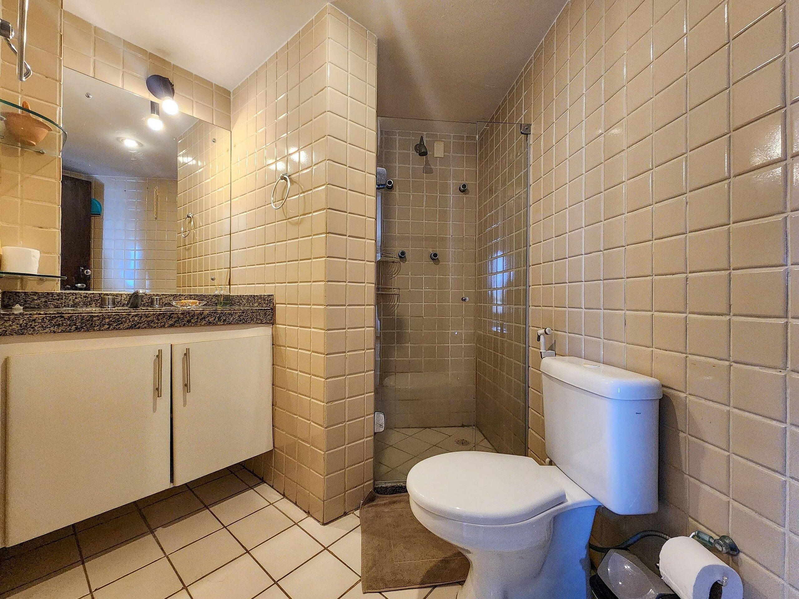 Une salle de bain avec des murs carrelés de couleur beige, des toilettes blanches, une douche en verre et un comptoir en granit avec un évier.