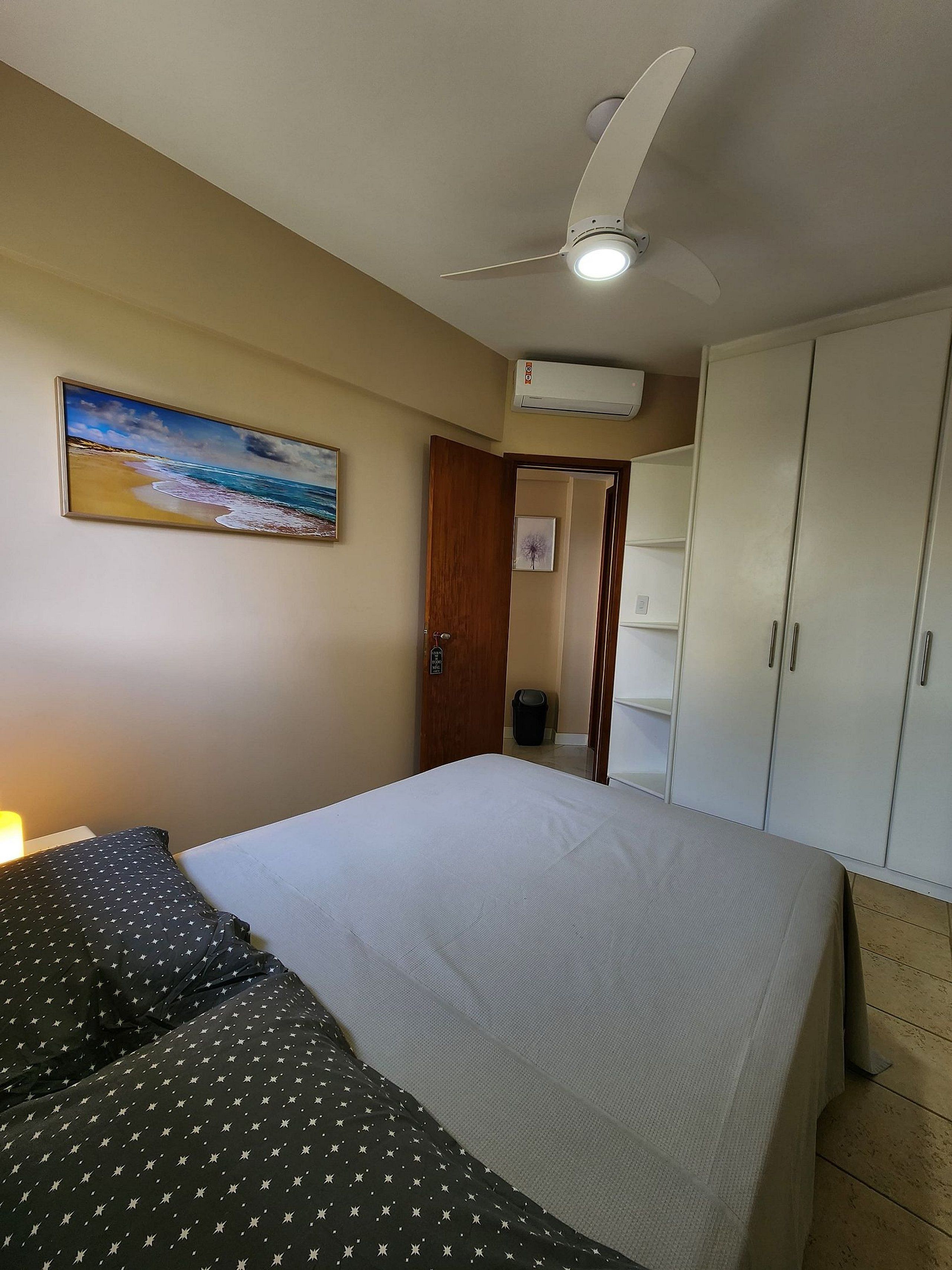 A bedroom with a bed, a ceiling fan, an air conditioning unit, a wardrobe, and a painting of a beach scene.