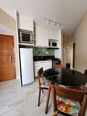 A compact kitchen with white cabinets, a microwave, a refrigerator, and a dining table with floral chairs. - Sol Nascente (Salvador)