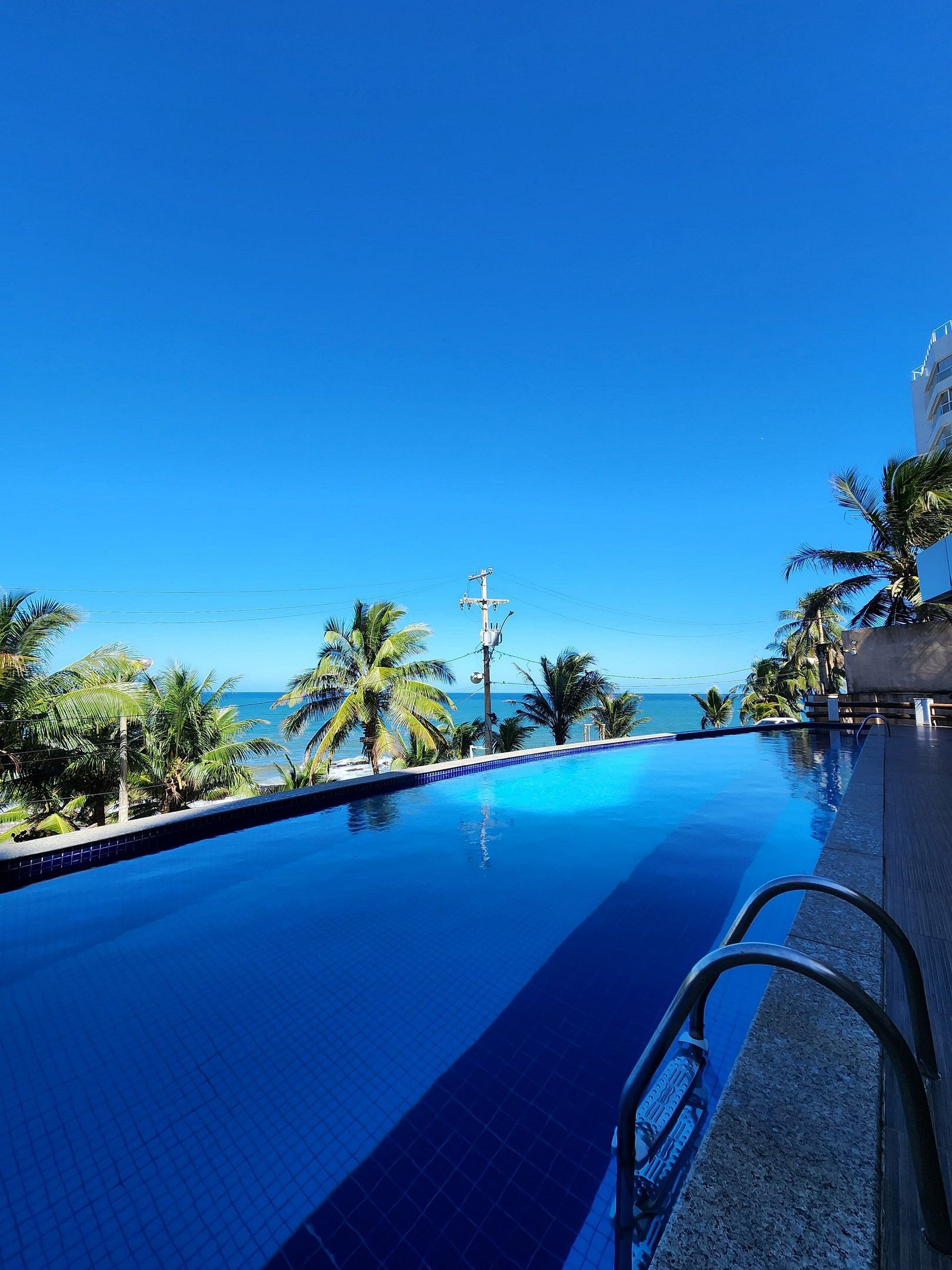Ein klares, blaues Schwimmbecken mit Blick auf den Ozean, umgeben von Palmen und einem Gebäude.